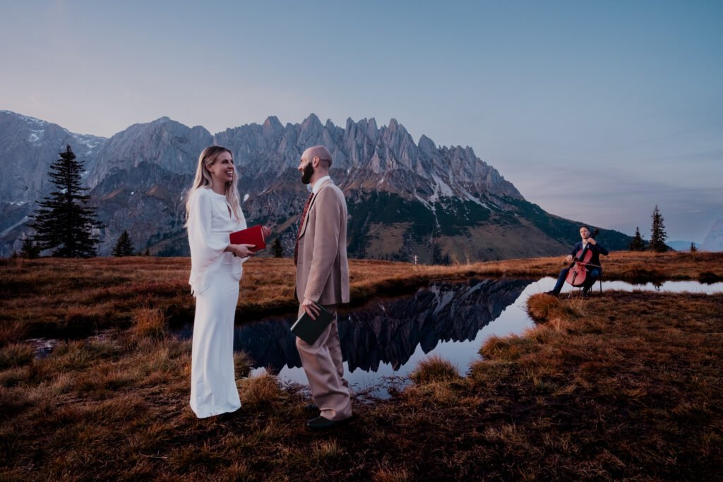 Couple holding vow books beside an alpine pond while a cellist plays nearby.
