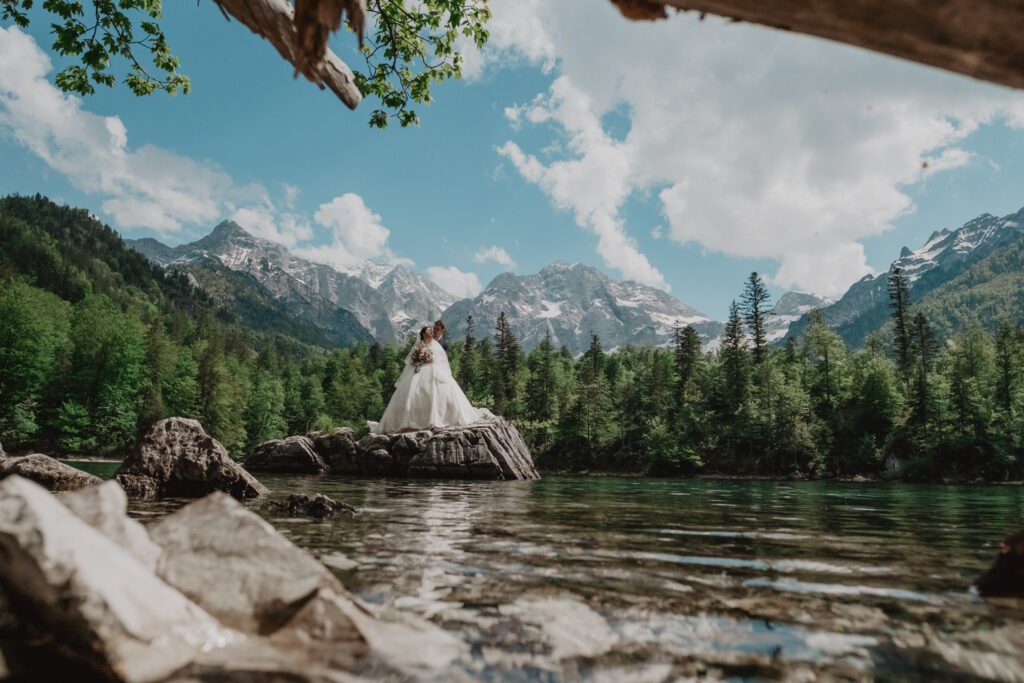 Couple in wedding attire standing on a rock in a clear mountain lake.