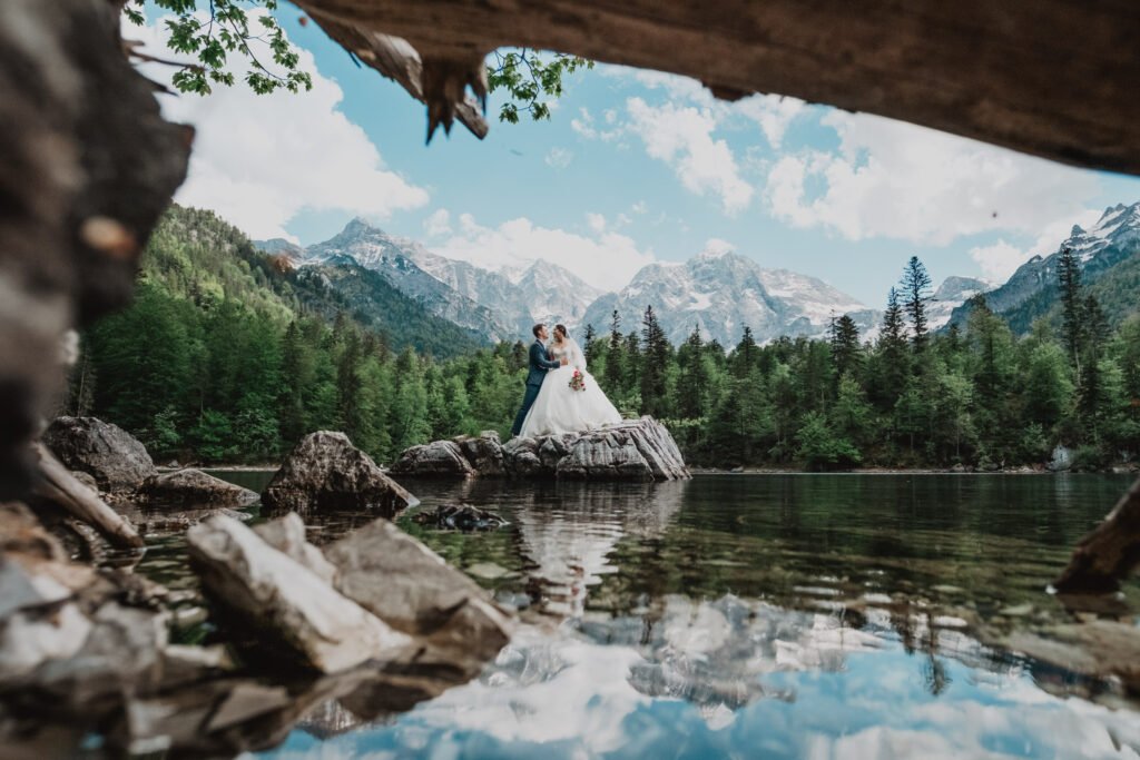 Bride standing on a rock reflected in a mountain lake, framed by branches.