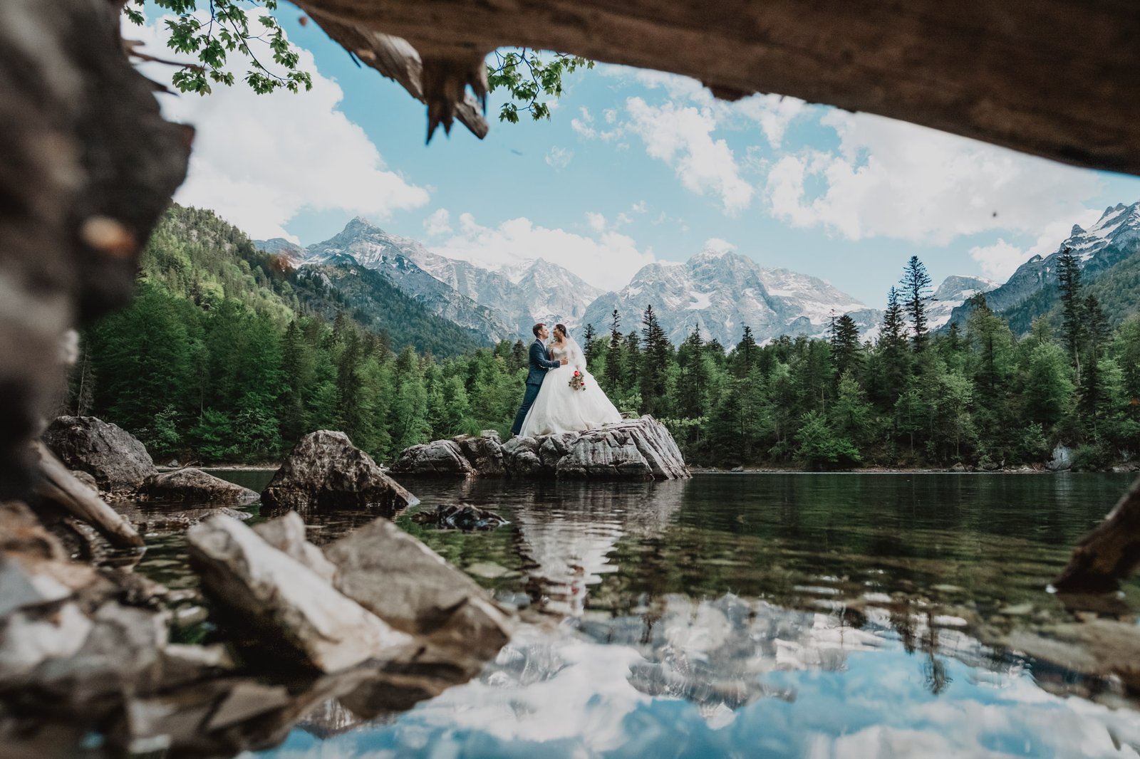 Braut auf einem Felsen spiegelt sich im Bergsee, eingerahmt von Zweigen.