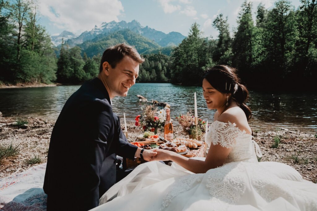 Couple holding hands beside a lakeside picnic, with forest and mountain peaks behind them.