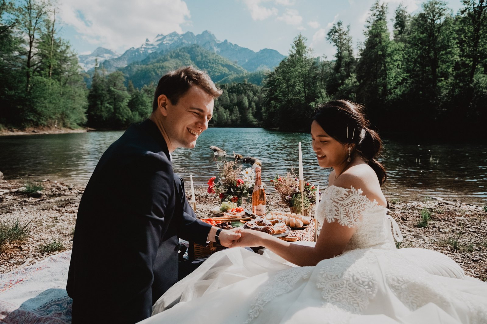 Paar hält sich neben einem Picknick am See an den Händen, mit Wald und Gipfeln im Hintergrund.