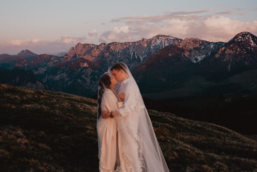 Couple embracing beneath a flowing veil with pink mountain peaks behind them.