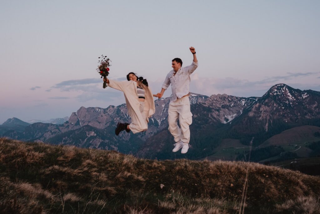 Couple jumping together on a mountain ridge, the bride holding her bouquet.