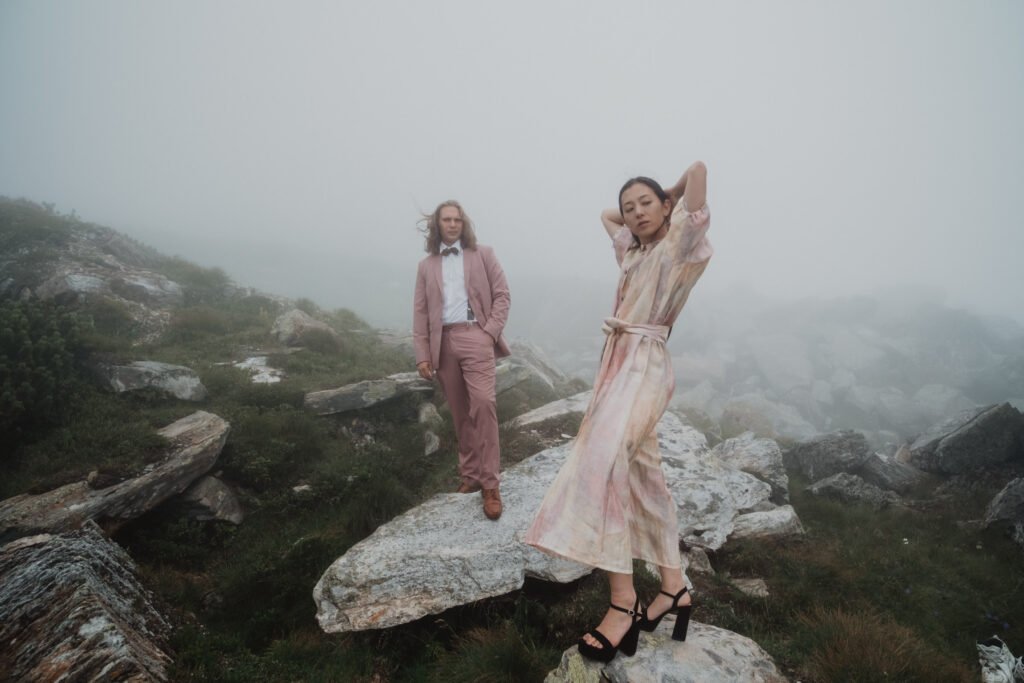 Bride lifting her veil while her partner watches on a misty mountain path.