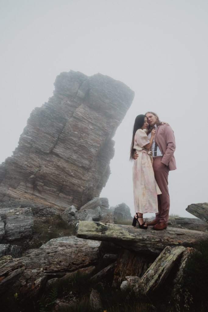 Couple leaning together beside a rock tower on a misty mountainside.