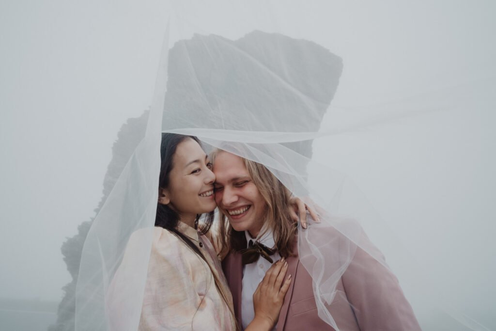 Couple laughing beneath a veil with a misty rock formation behind them.