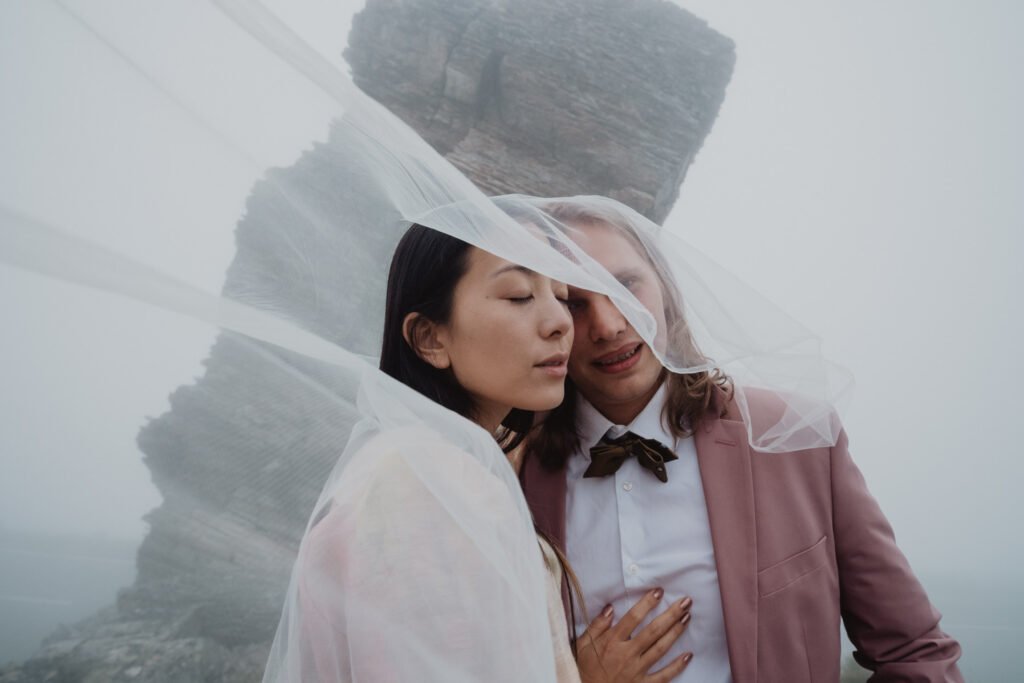 Couple beneath a veil in the mist beside a rocky mountain formation.