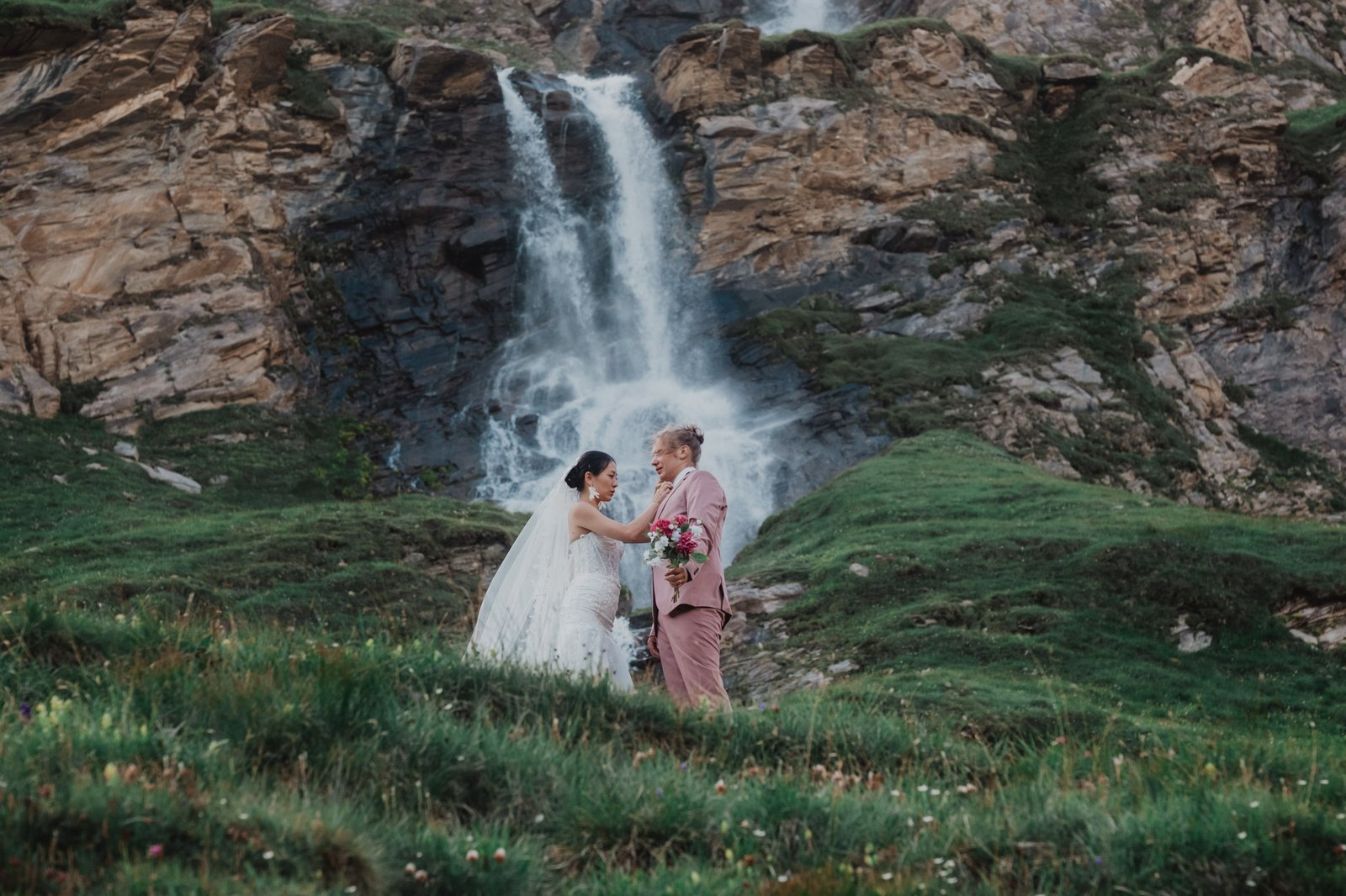 Paar unter einem Wasserfall an einem grünen Berghang.