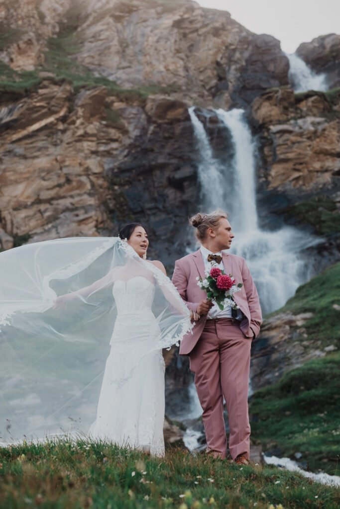 Couple holding hands with the bride's veil flowing beside a waterfall.