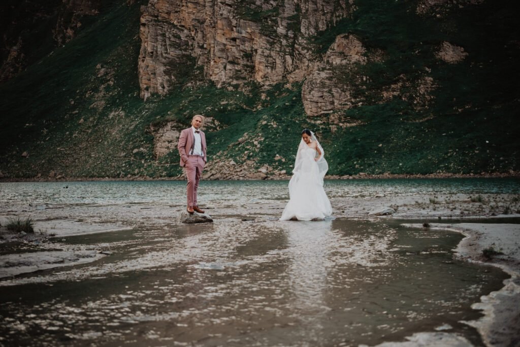 Couple standing apart on a rocky lakeshore beneath a steep mountainside.