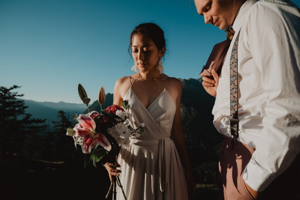 Bride holding a bouquet beside her partner on a sunlit mountain slope.