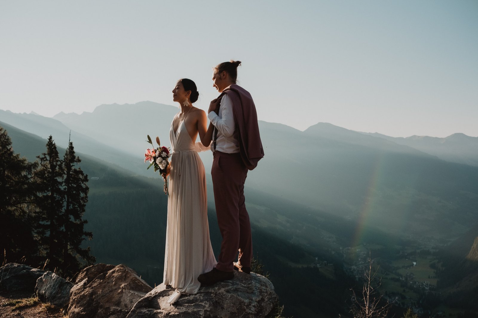 Couple in wedding attire looking across a mountain valley from a rocky viewpoint.