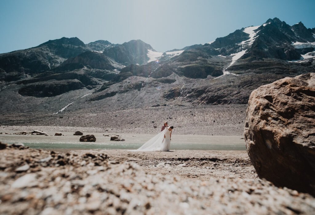 Wide view of a couple beside a lake beneath a snowy mountain massif.