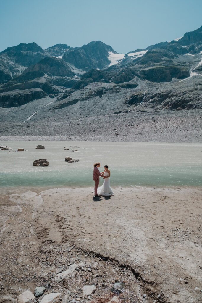 Couple standing beside a glacier lake beneath snowy mountain peaks.