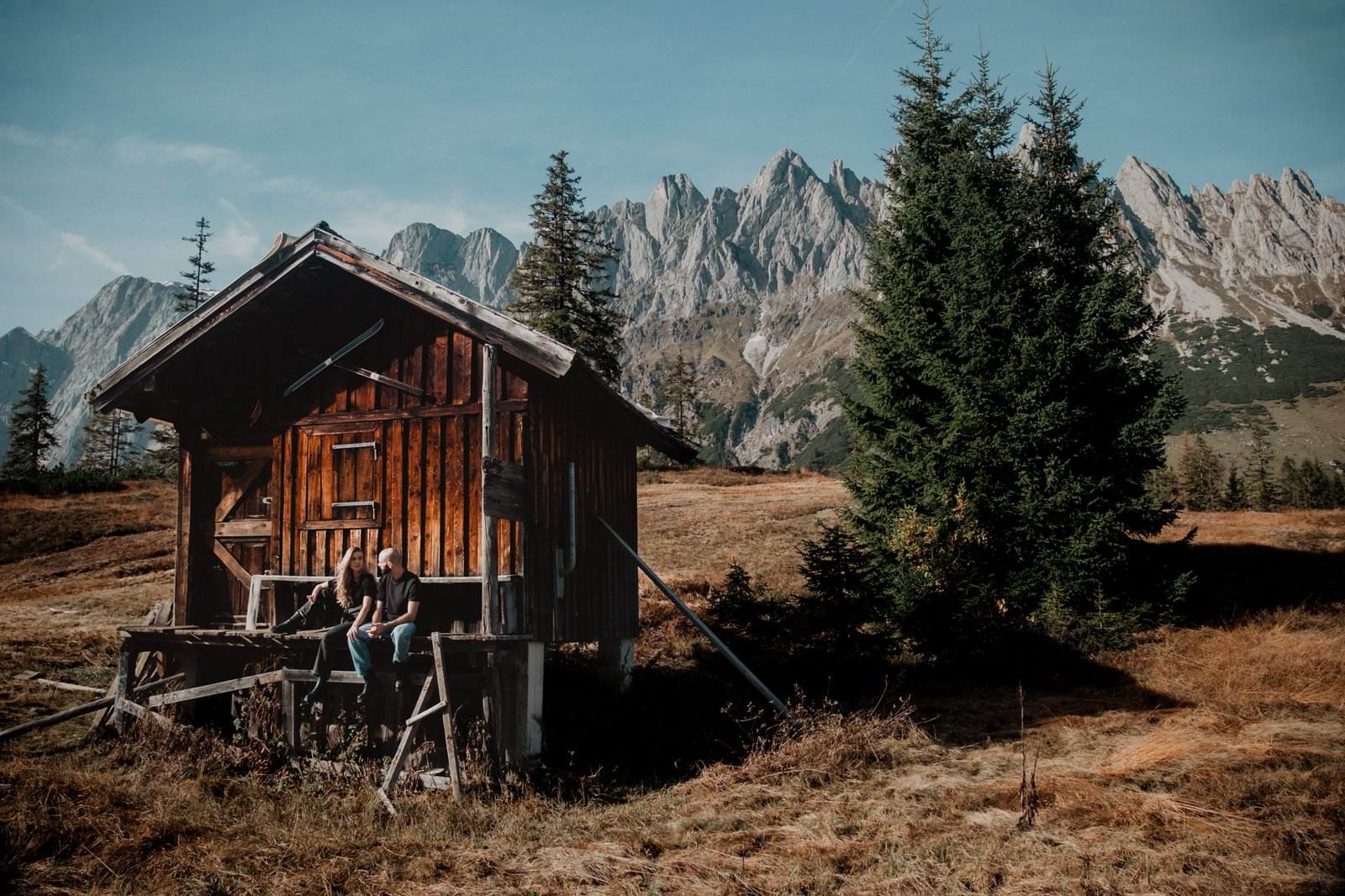 Couple sitting outside a wooden alpine hut beneath jagged mountain peaks.