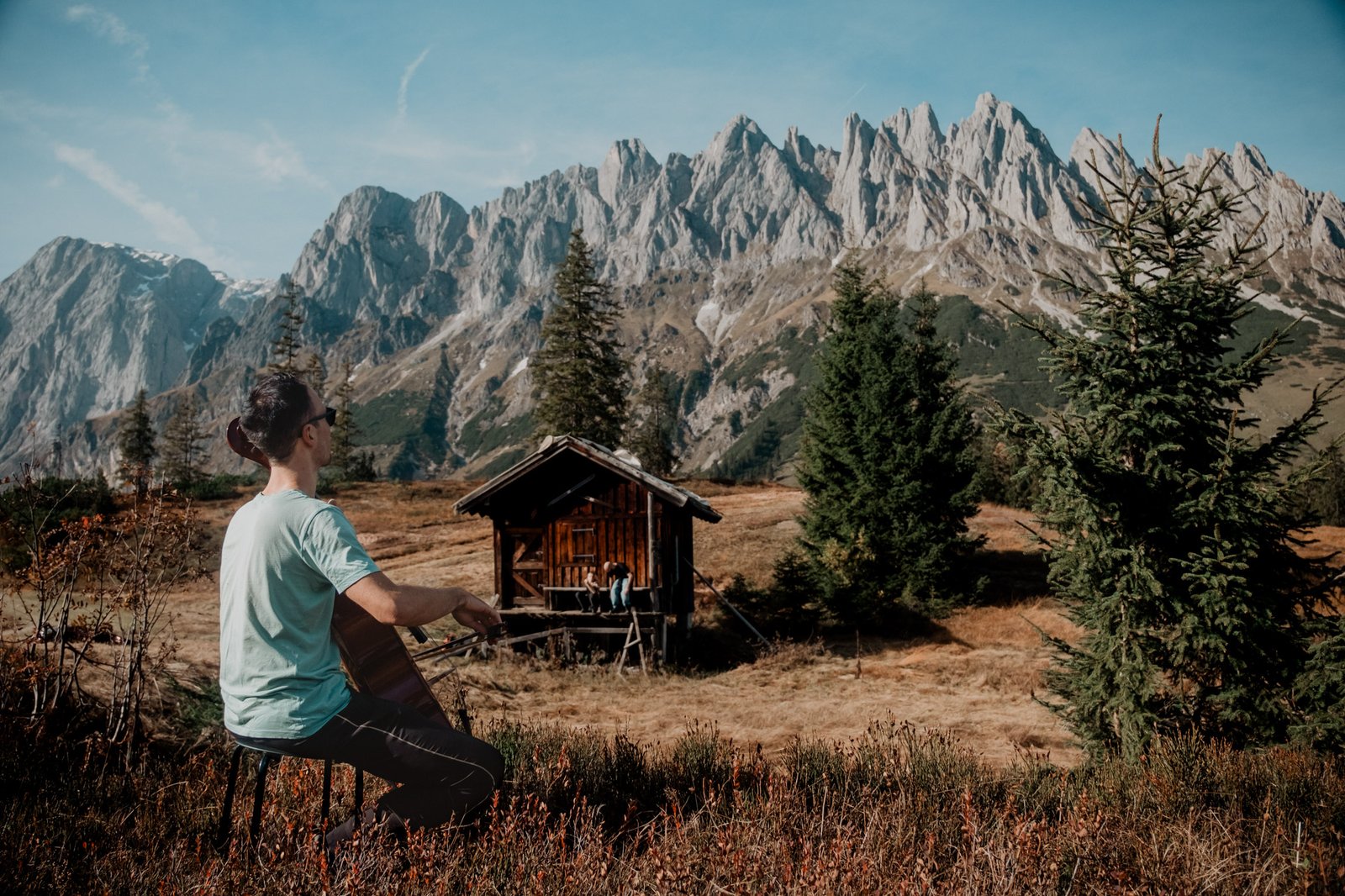 Mann zeigt neben einer Holzhütte auf die Berglandschaft.