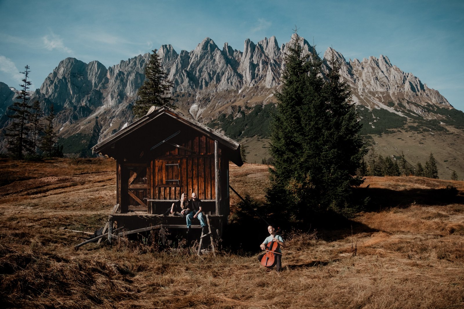Cellist neben einer Berghütte, auf deren Veranda ein Paar sitzt.