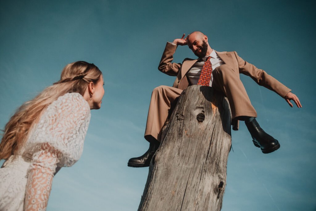 Groom perched on a tall tree stump while the bride laughs beside him.