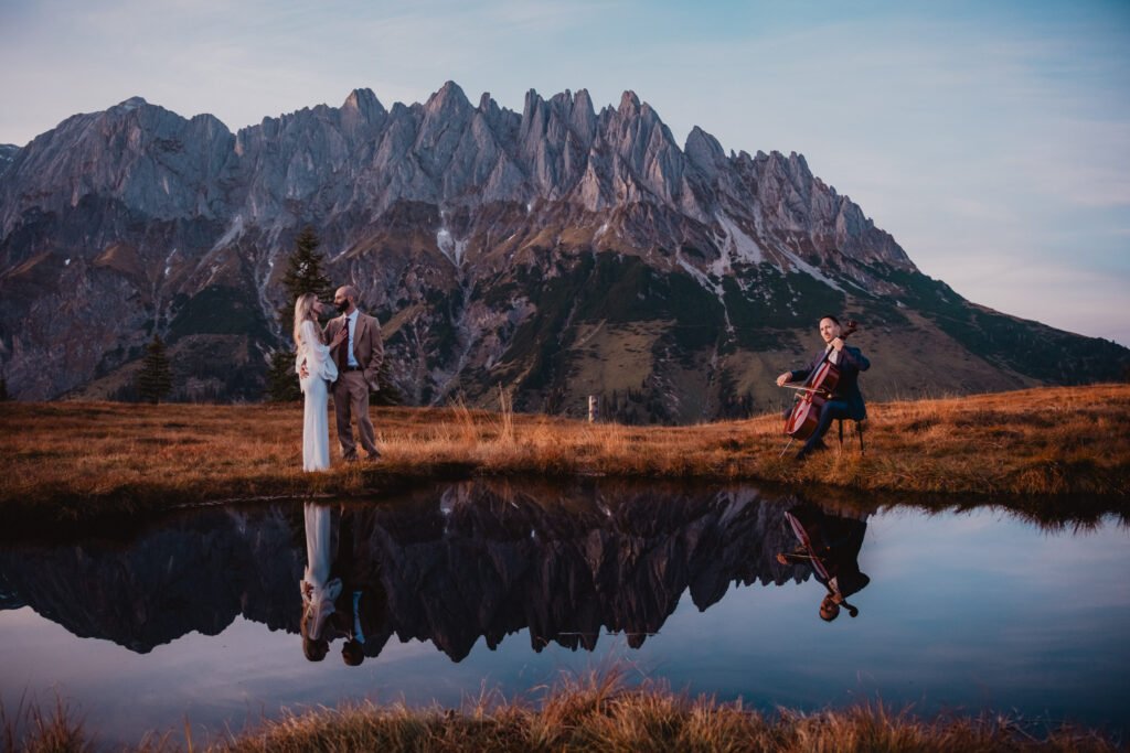 Cellist accompanying a couple beside a mountain pool at sunset.