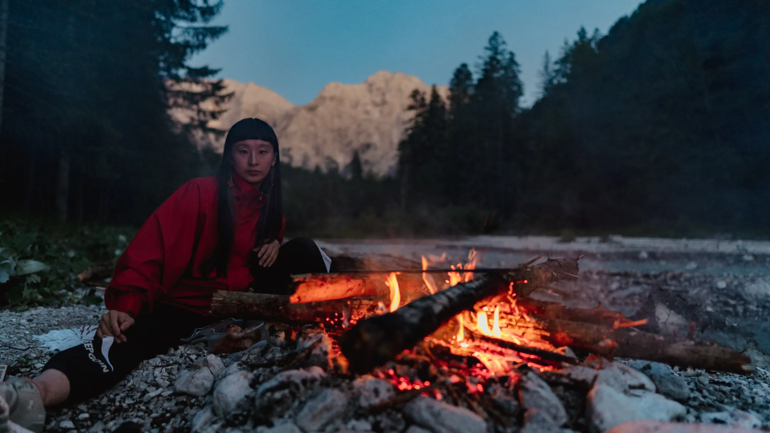 Frau sitzt an einem Lagerfeuer vor Wald und Bergen.