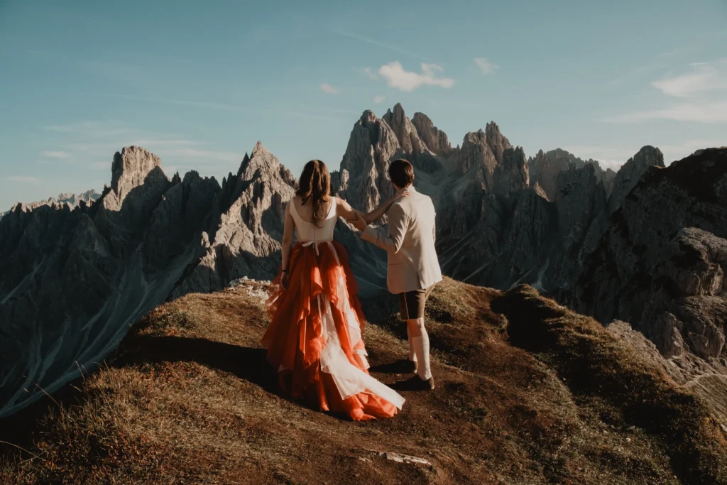 Couple facing each other on a grassy ridge with dramatic mountains behind them.