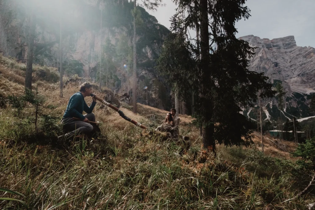 Gregor playing a didgeridoo beside a tree in an alpine meadow.