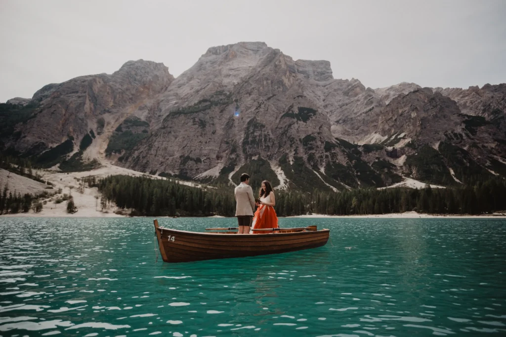 Couple standing in a wooden rowing boat on a turquoise lake beneath mountain cliffs.