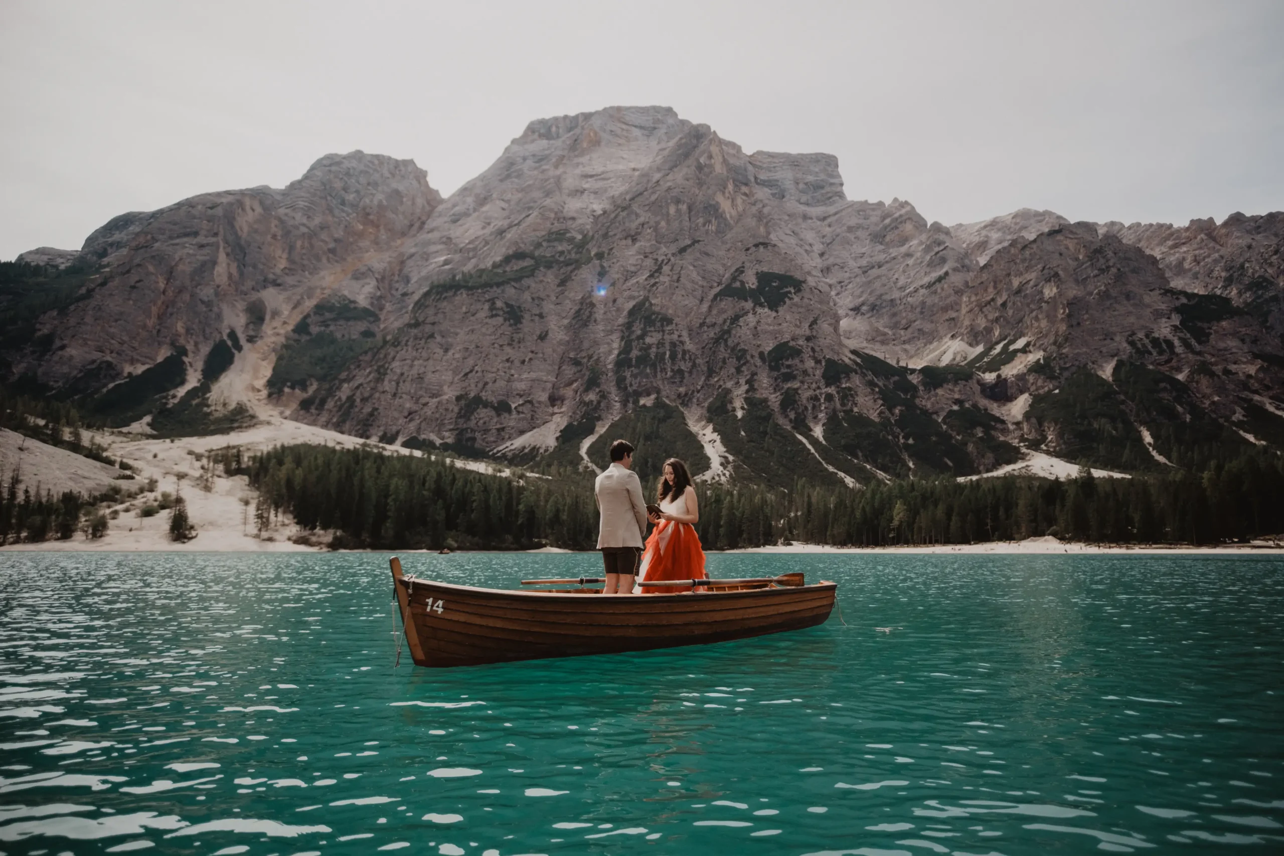 Couple standing in a wooden rowing boat on a turquoise lake beneath mountain cliffs.