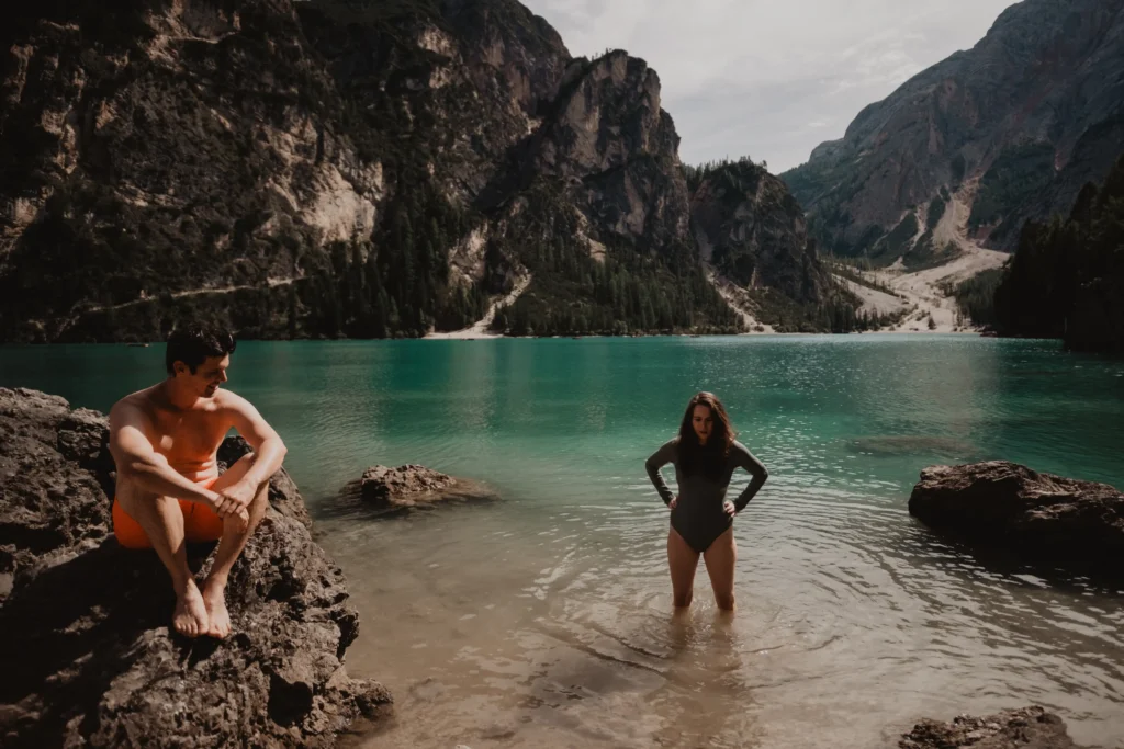 Couple in swimwear at the lake, one in the water and one seated on the shore.