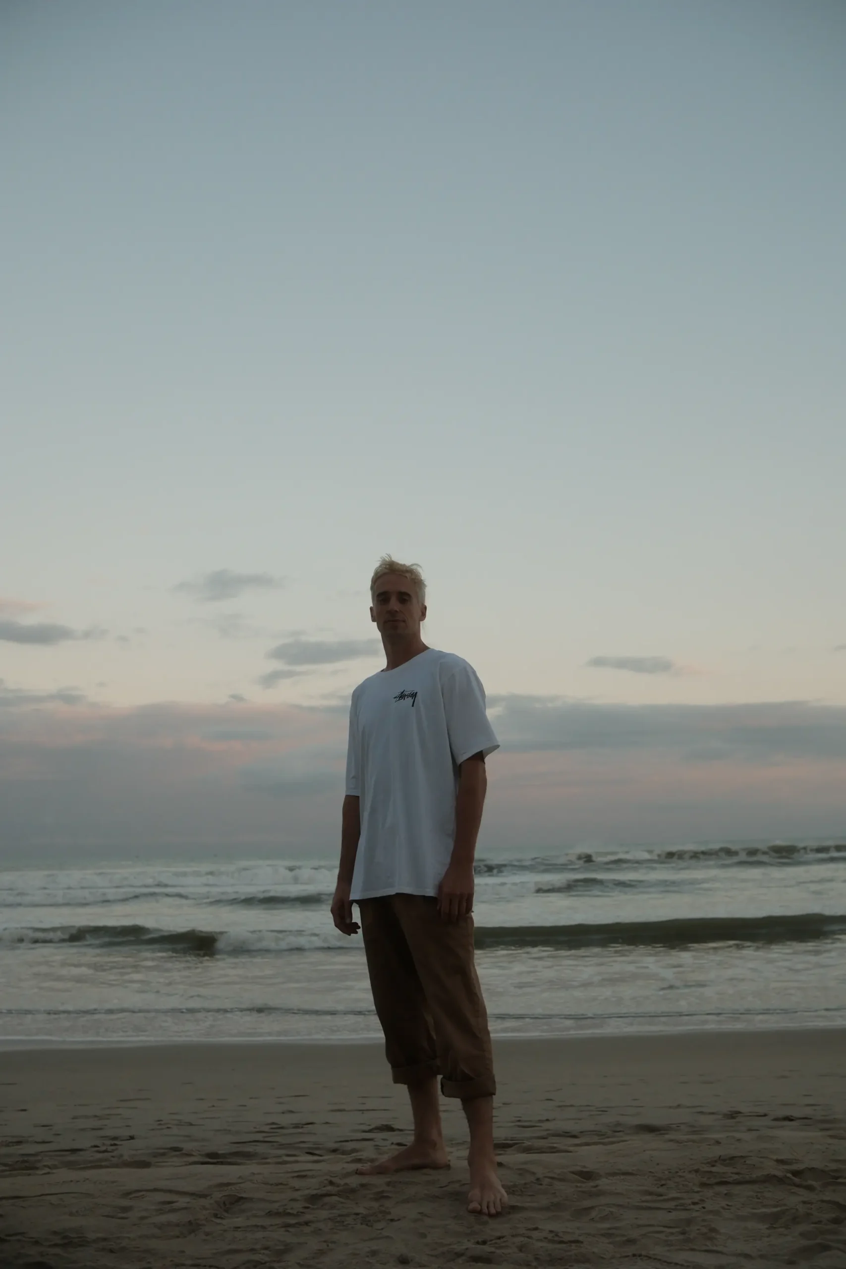 Elias steht in der Dämmerung barfuß am Strand.