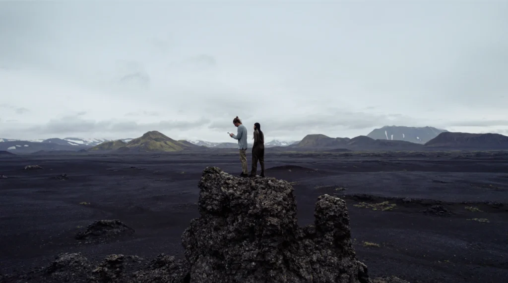Two figures standing on a rocky outcrop in a wide volcanic landscape.
