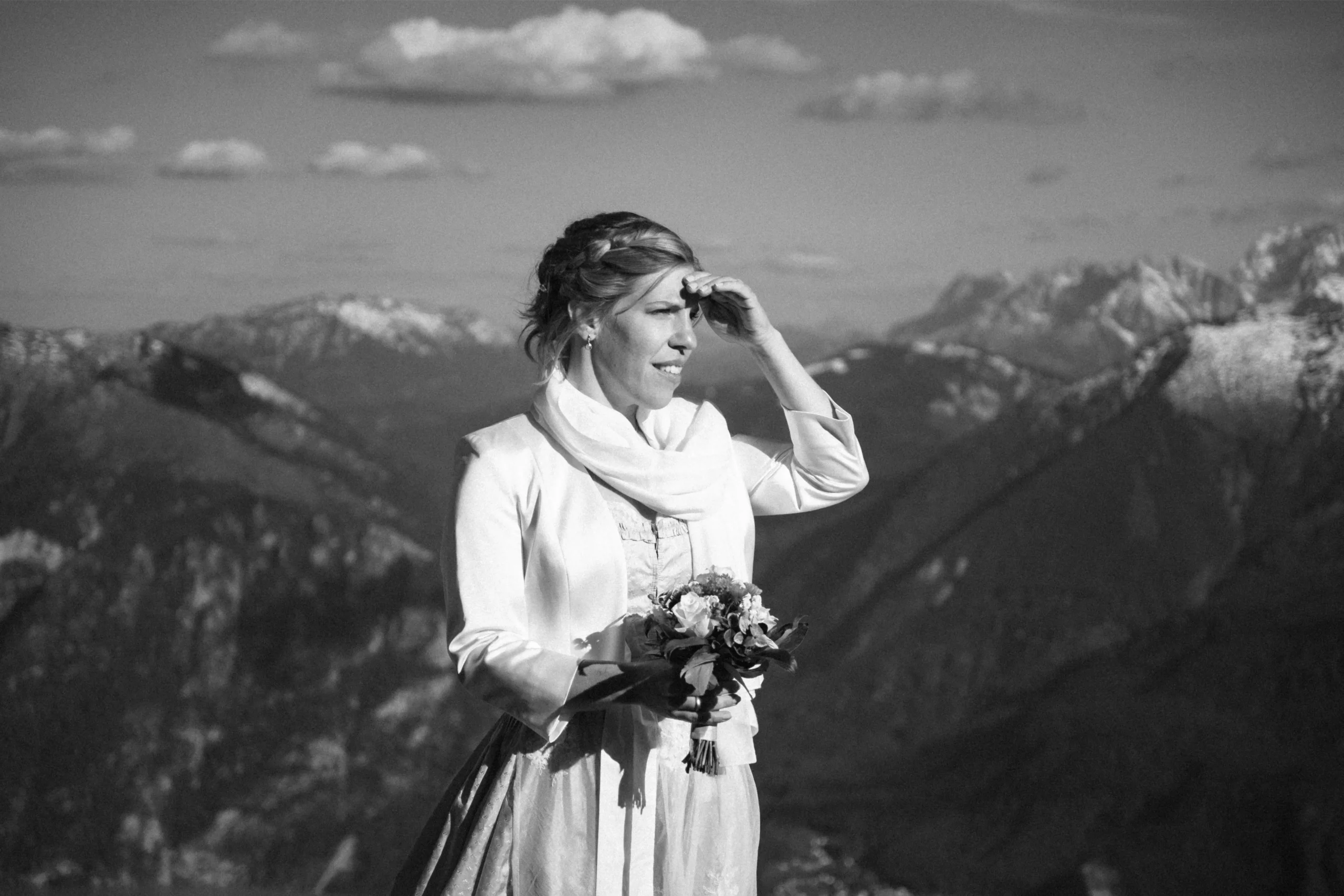 Black-and-white portrait of a bride holding flowers and looking towards snowy mountains.