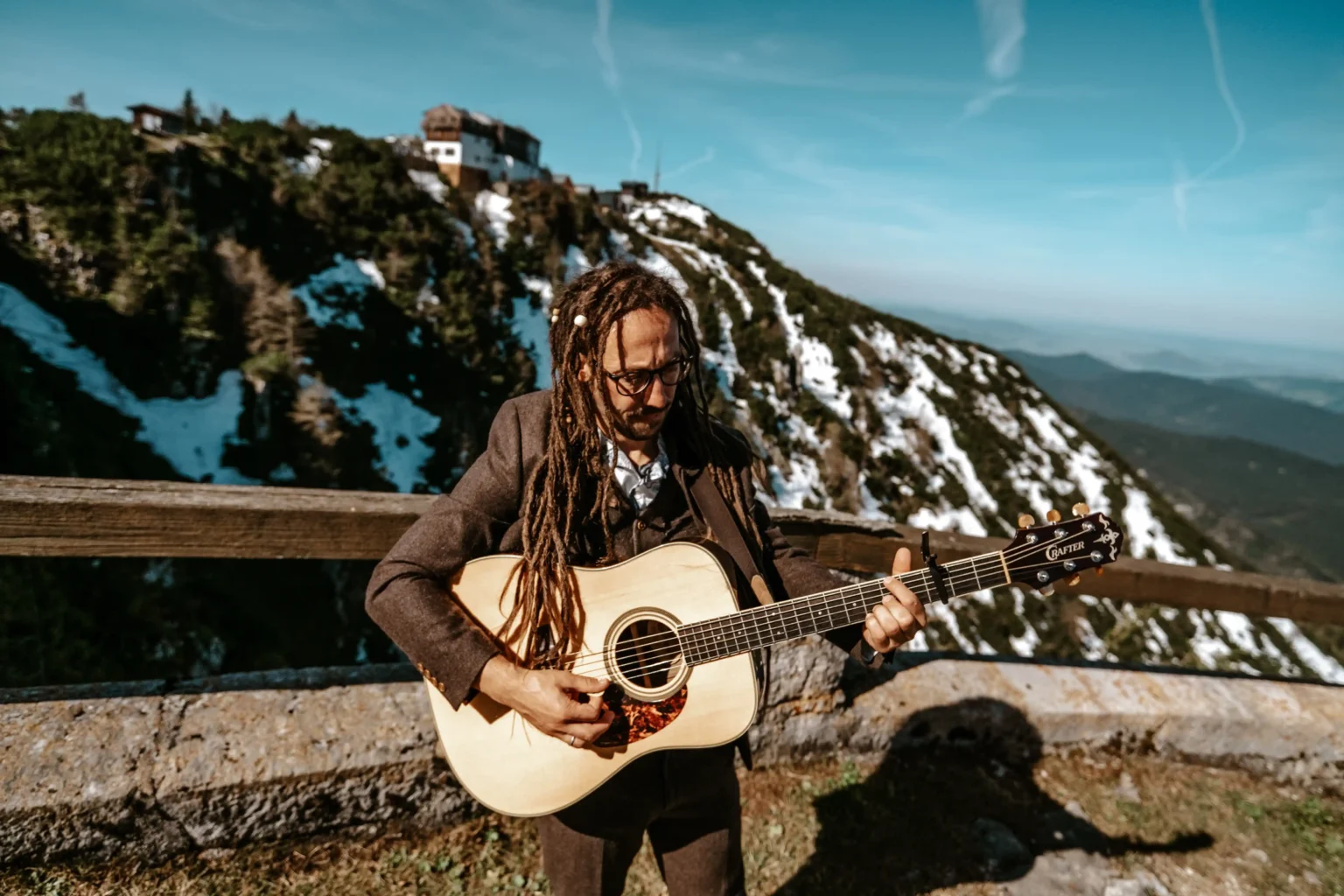 Beda mit Palme playing acoustic guitar on a wooden terrace with mountain scenery behind him.