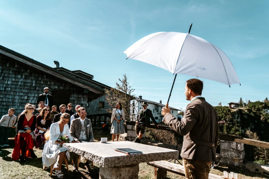 Groom holding a white umbrella during the ceremony with guests behind him.