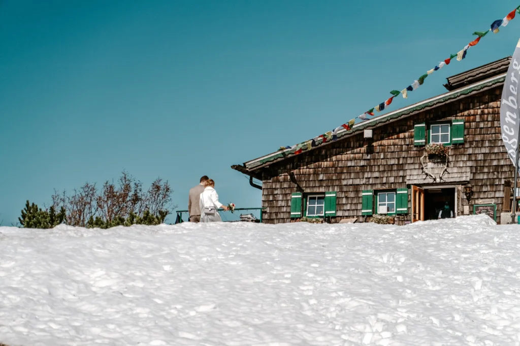 Mountain hut with snow in the foreground and people beside it.