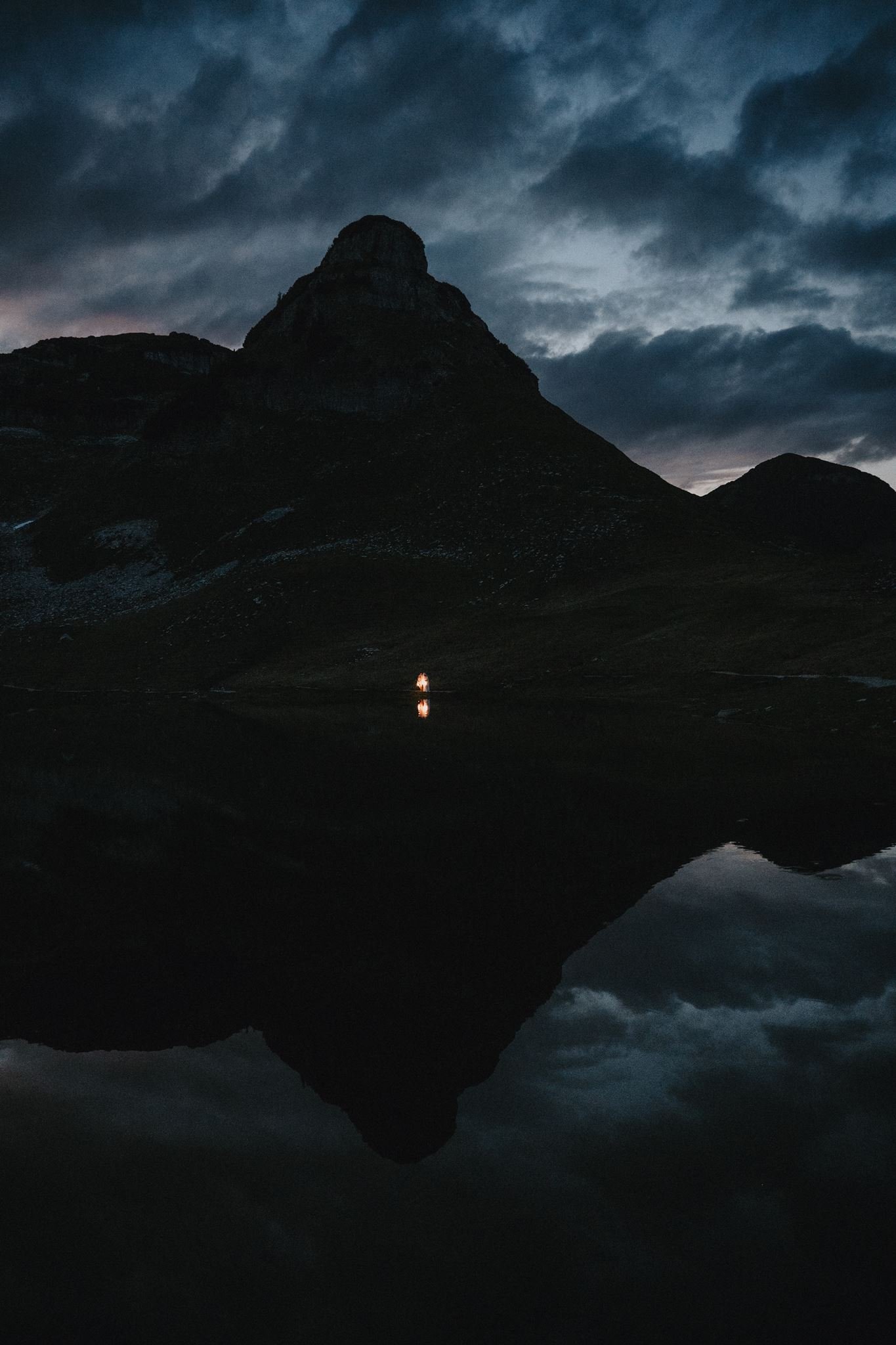 Bergsilhouette und kleines Licht spiegeln sich nachts in einem See.