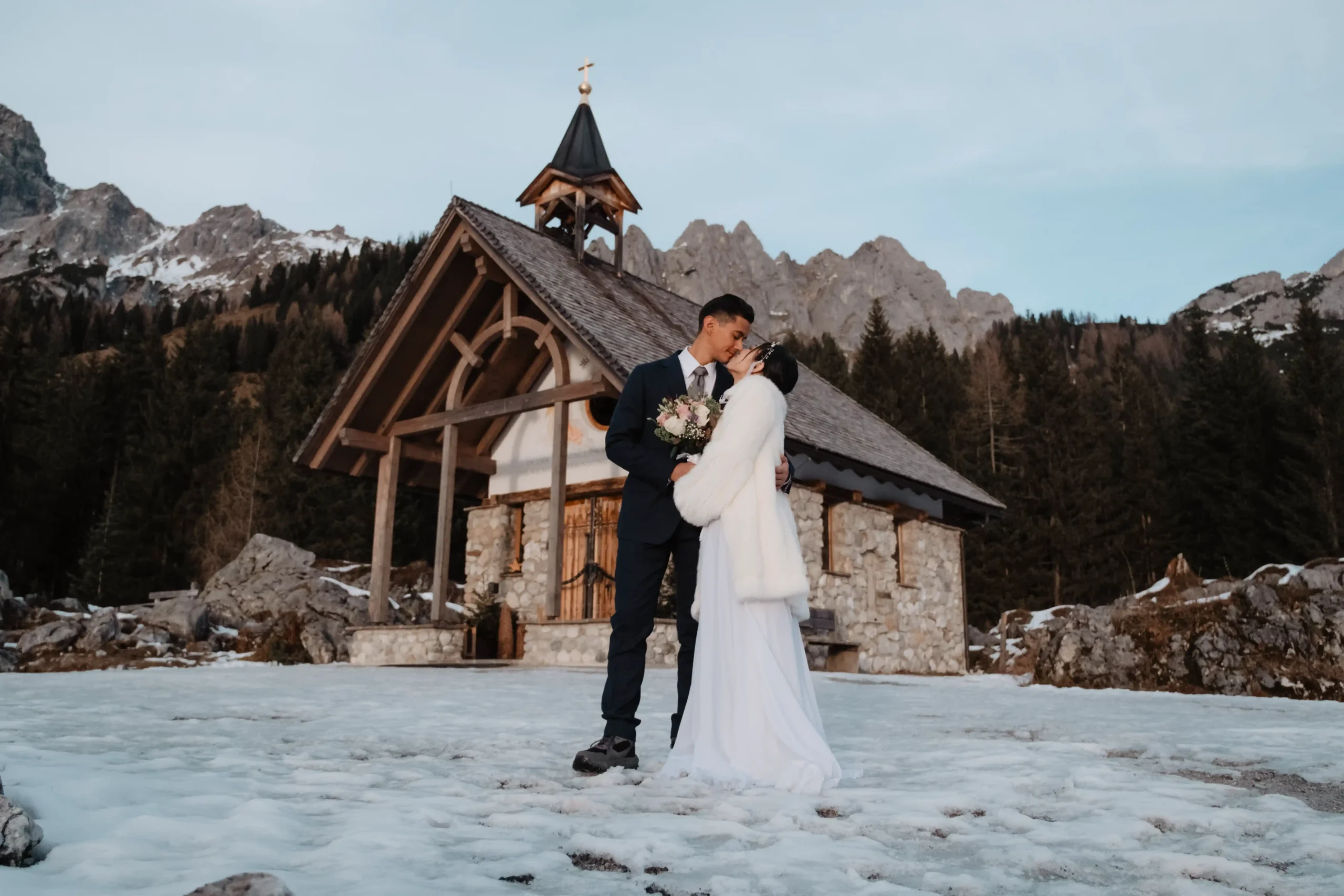 Couple embracing in the snow outside a small mountain chapel.
