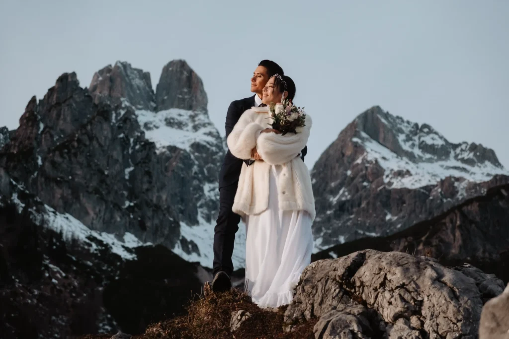 Couple embracing on a rocky viewpoint with snow-covered peaks behind them.