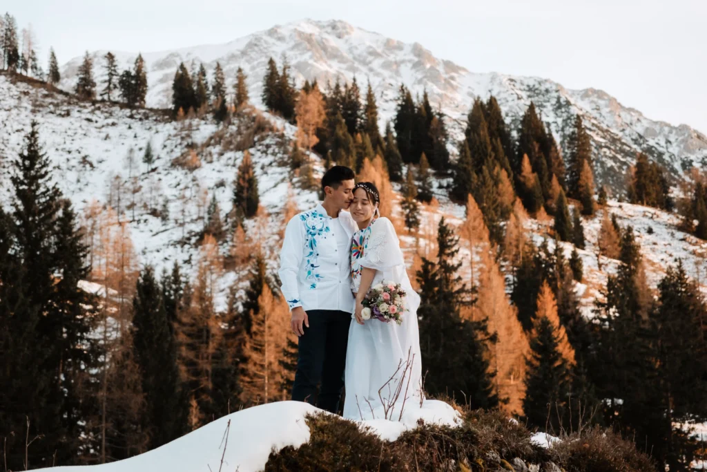 Couple in embroidered wedding attire standing among snow and golden larch trees.