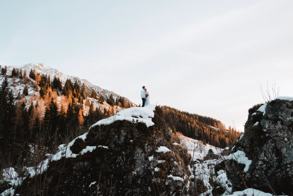 Couple standing on a distant snowy outcrop above an autumn forest.