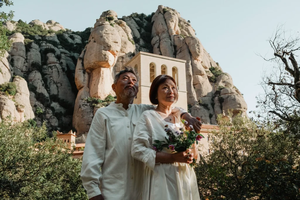 Couple with a bouquet beside a small chapel beneath rocky peaks.