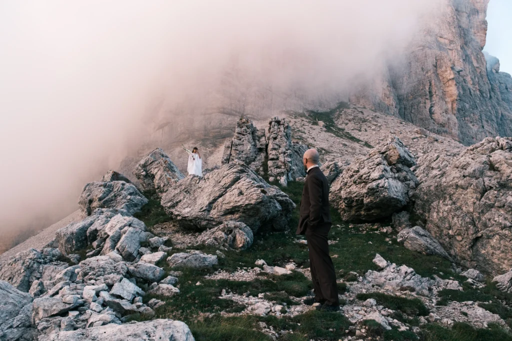 Groom standing on a rocky path with the bride farther along the misty ridge.