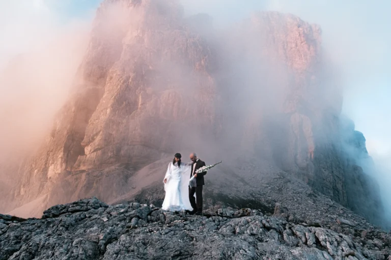 Couple in wedding attire standing on a rocky ridge beneath a mist-covered Dolomites cliff.