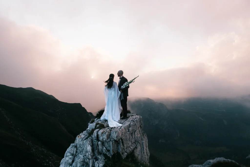 Couple standing on a rock pinnacle beneath pink evening clouds.
