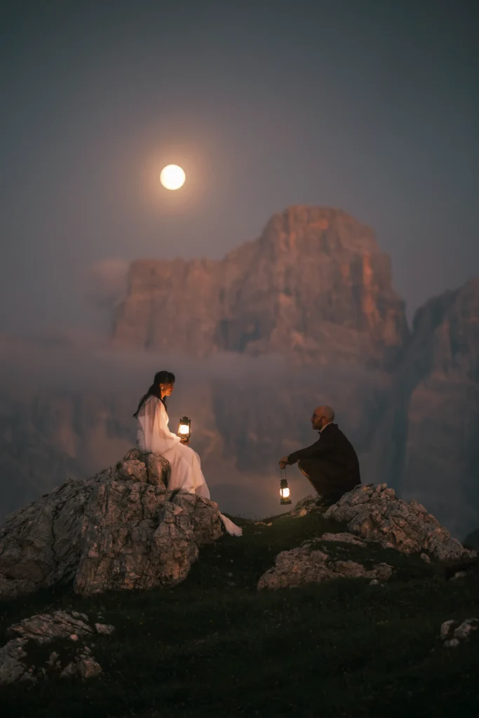Couple sitting opposite each other with lanterns beneath a moonlit mountain peak.