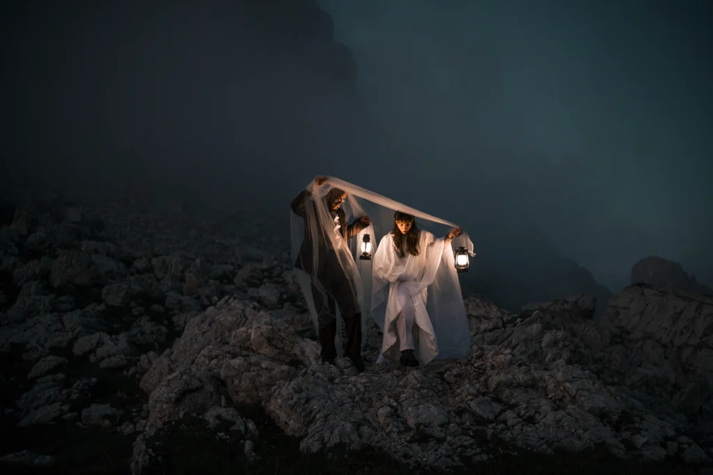 Couple holding lanterns beneath a veil on a rocky mountain path at dusk.
