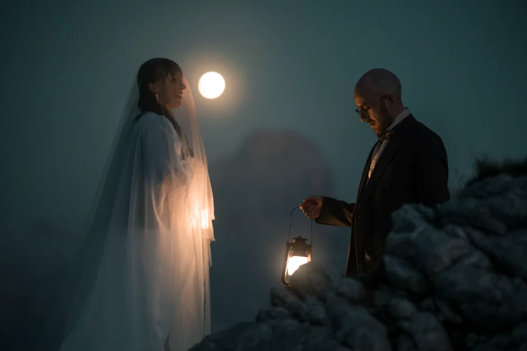 Bride and groom facing each other beside a lantern on a rocky ridge at night.