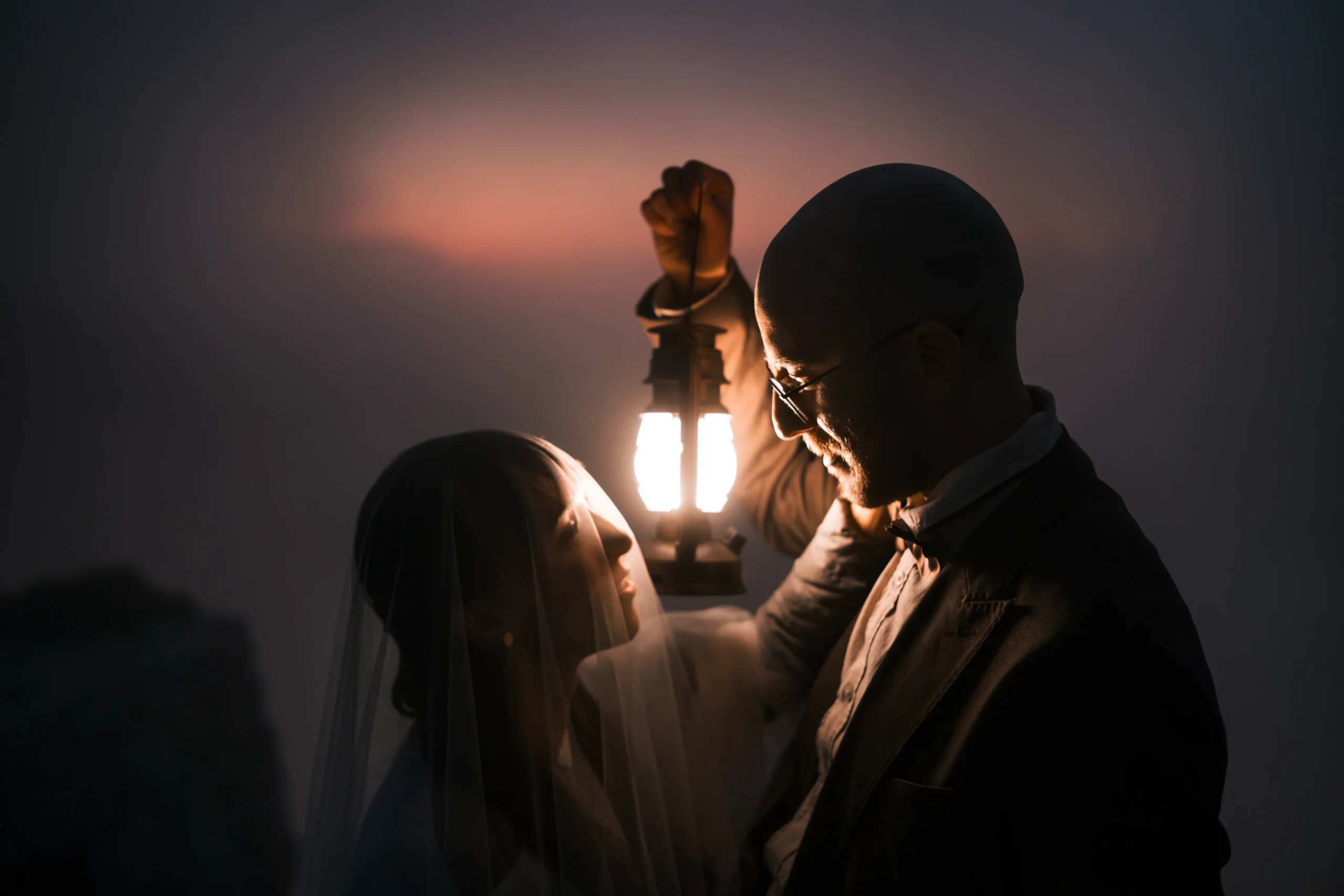 Couple smiling at each other in the glow of a lantern at dusk.