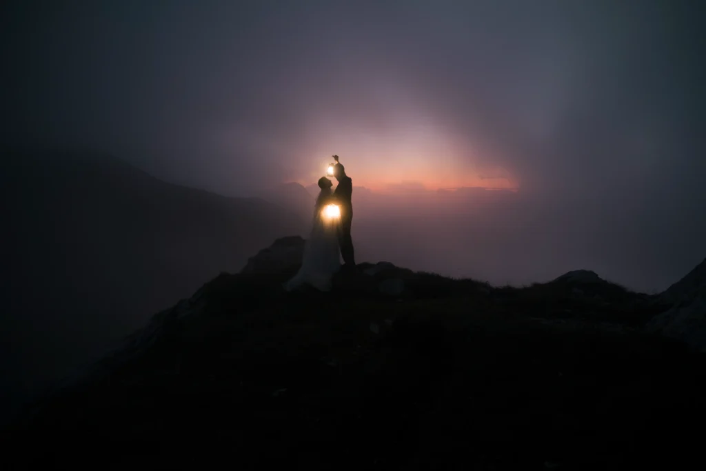 Couple silhouetted with glowing lanterns on a mountain ridge at dusk.
